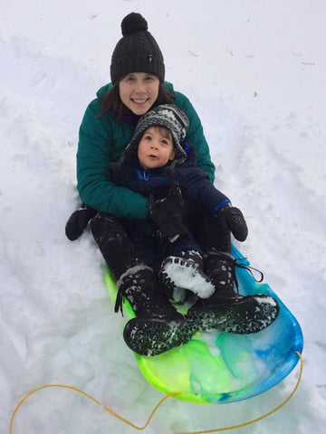 A woman holding a young child on a sled while wearing a Smart Wool had, Arcteryx jacket, and Columbia Snow Boots