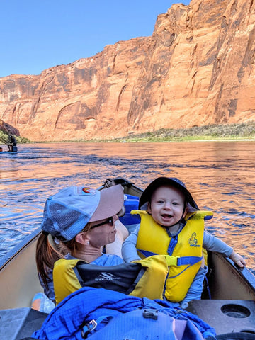 A woman holding a baby with a life jacket on in a canoe while floating on a river in a canyon