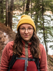 A woman hiking in a forested area wearing a yellow beanie, glasses, and a pink sweater