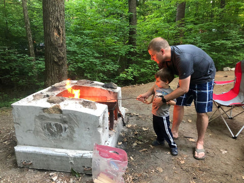 A man helping a little boy roast a marshmallow over a fire pit at a campground