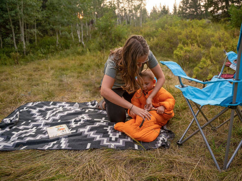 Woman zipping a small child into a Morrison Outdoors Big Mo sleeping bag in an outdoor setting