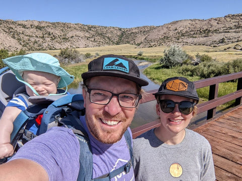 A woman with a man wearing a baby in a Kelty hiking carrier walking across a bridge with a mountainous view 