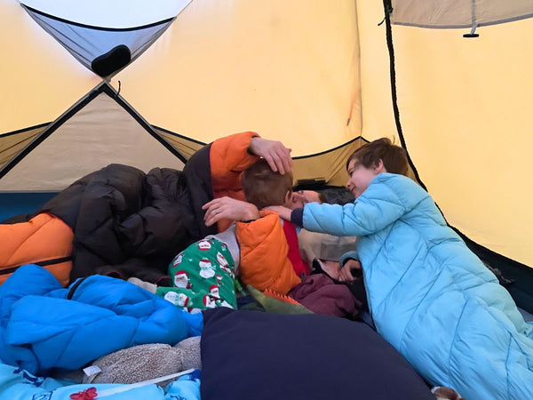 A man playing with his sons inside a tent while wearing a Morrison Outdoors sleeping bag and a Horizon Hound down blanket