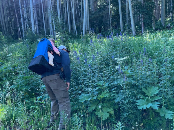 A man wearing a baby in a framed carrier on his back while they explore a field of wildflowers