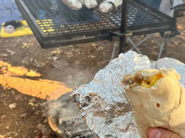 A breakfast burrito being eaten with other burritos on a camp grill in the background