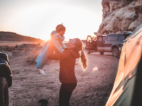 A woman holding a small child wearing a Morrison Outdoors sleeping bag near a campsite