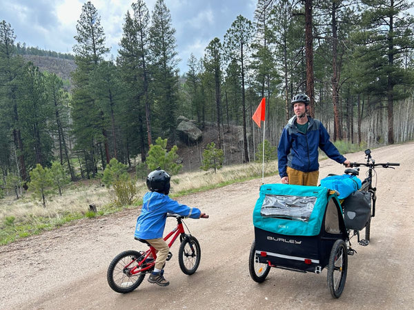 A young child on a red bike and A man standing next to a bike with a Burley trailer attached