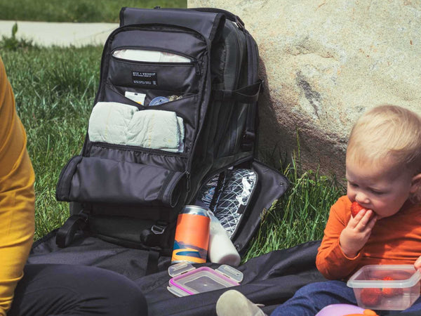 A baby eating while sitting on a mat outdoors next to an opened Milk X Whiskey Adventure Proof Diaper Backpack