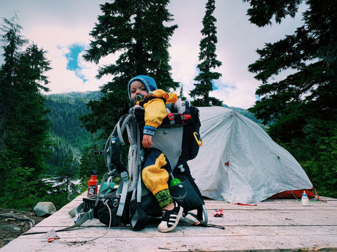 A toddler in an Osprey Poco hiking pack in front of a tent on top of a camping platform