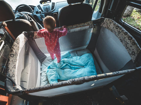 A baby standing inside a pack and play inside a van in a forested area.